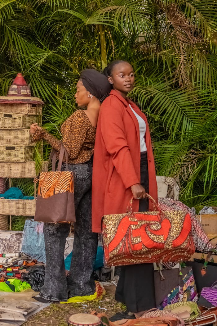 Two stylish women showcasing colorful bags at an outdoor African market.